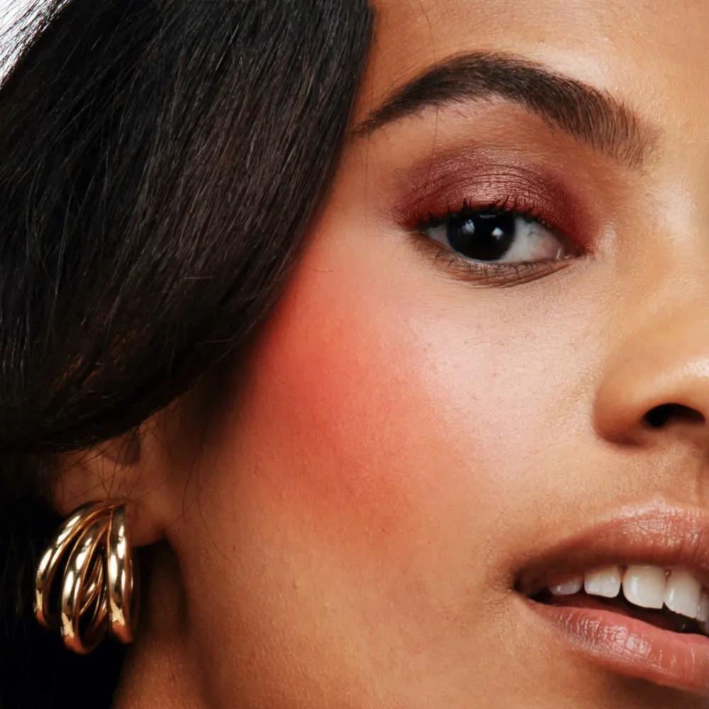 Close-up of a woman's face featuring smooth, matte, natural pink blush on cheeks with subtle eye makeup and gold hoop.